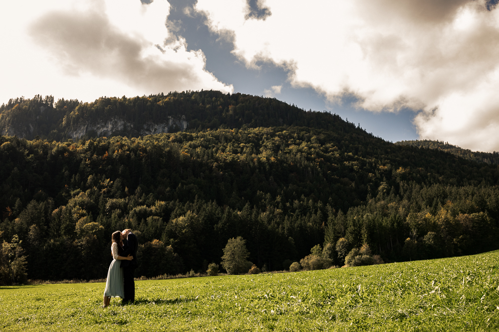 Hochzeitspaar steht auf Wiese vor kleinem Berg und küsst sich